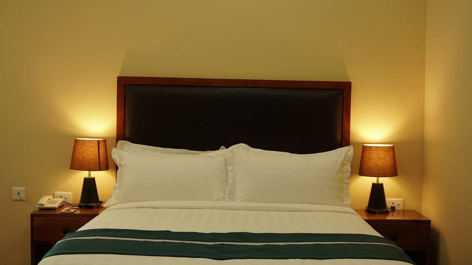 A double room with bedside lamps lit either side of a padded headboard and a green runner across white linen