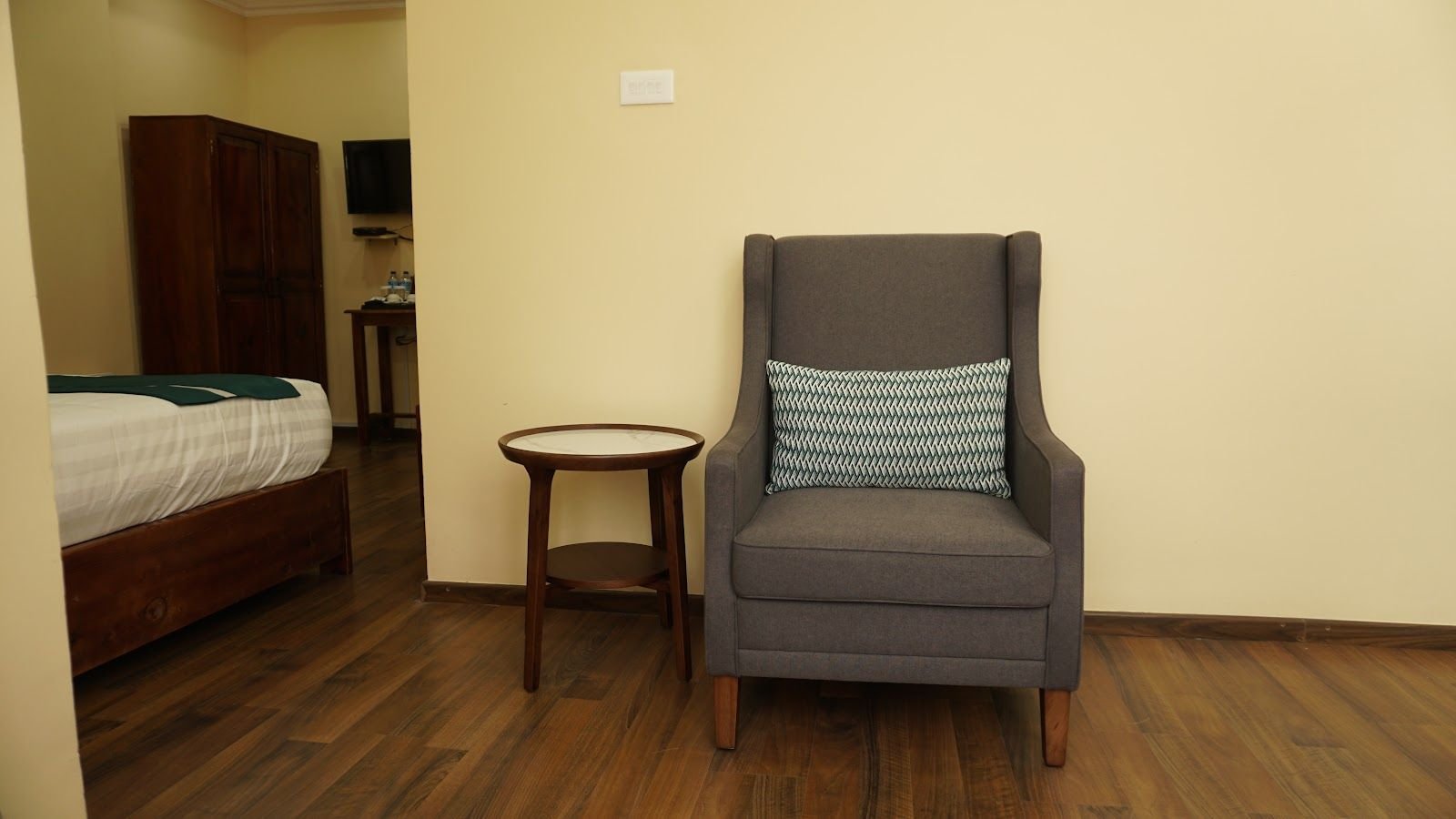 A grey wing-back armchair and small round side table beside the bed