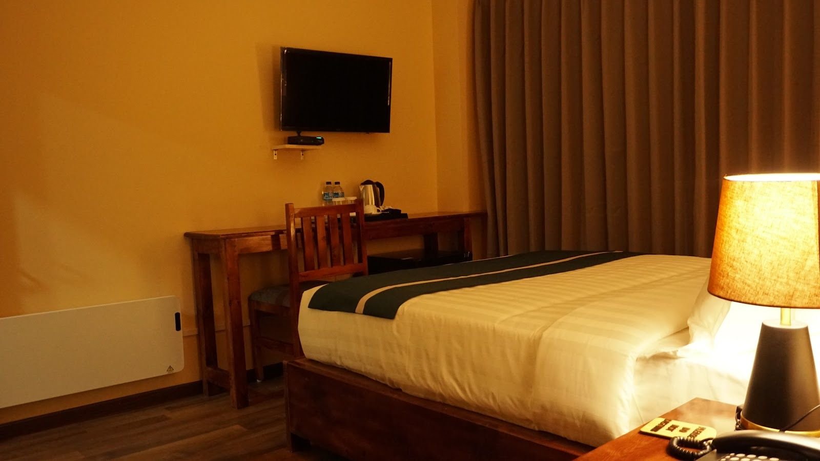 A guest room with a wall-mounted television, writing desk and a lamp beside the bed